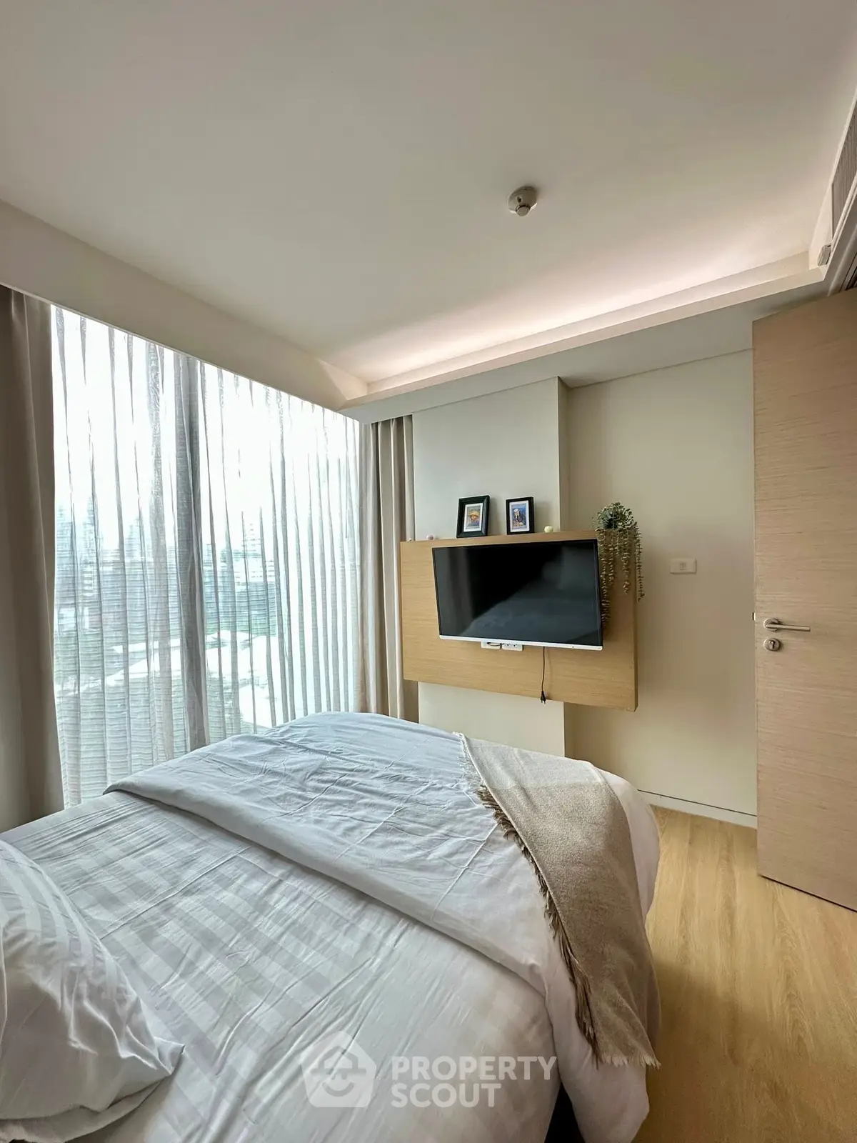 Modern bedroom with natural light and sleek design, featuring a mounted TV and cozy bedding.