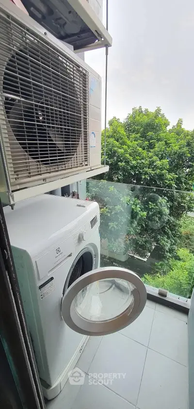 Compact balcony with washing machine and air conditioning unit, overlooking lush greenery.