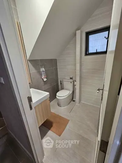 Modern bathroom with sleek design under sloped ceiling, featuring compact fixtures and neutral tones.