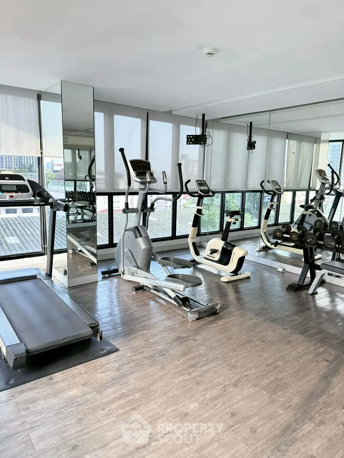 Modern gym with cardio equipment and weights in a high-rise building with city views.