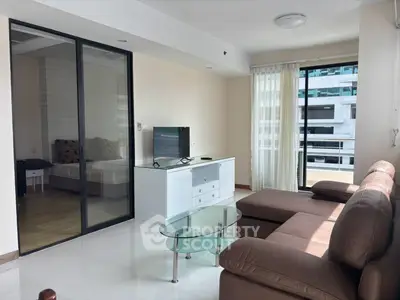 Modern living room with glass coffee table and brown sofa, adjacent to a bedroom with sliding glass doors.