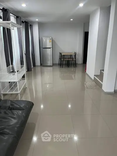 Spacious modern living area with sleek tile flooring and minimalist decor, featuring a dining table and refrigerator.
