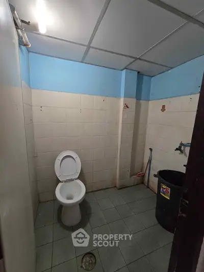 Basic bathroom with tiled walls and floor, featuring a toilet and cleaning supplies.
