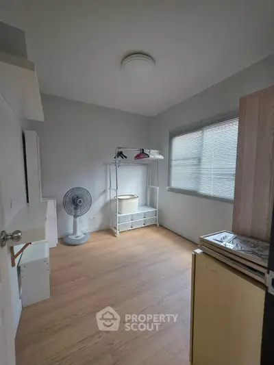 Cozy bedroom with wooden flooring and ample natural light, featuring a minimalist wardrobe and a standing fan.