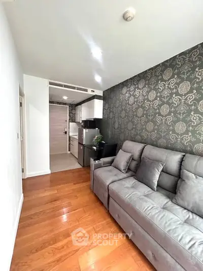 Modern living room with elegant wallpaper and cozy gray sofa, featuring sleek wooden flooring and compact kitchen area.