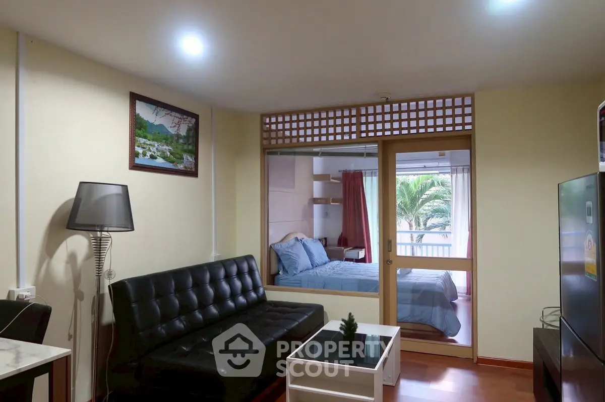 Cozy living room with black sofa and view into a bright bedroom with balcony access.