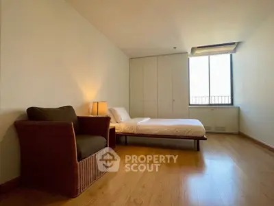 Spacious bedroom with wooden flooring and cozy wicker chair, featuring a large window for natural light.