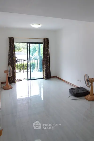 Spacious living room with sliding glass doors leading to a garden view, featuring elegant curtains and floor fans.