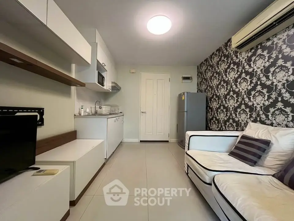 Modern living room with stylish wallpaper and compact kitchen area, featuring sleek furniture and air conditioning.