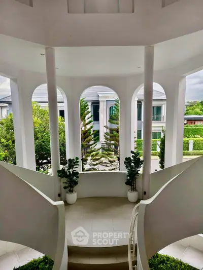 Elegant curved staircase with lush garden view through arched windows in luxury home.