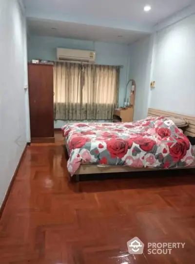 Cozy bedroom with floral bedding and wooden flooring, featuring an air conditioner and ample natural light.