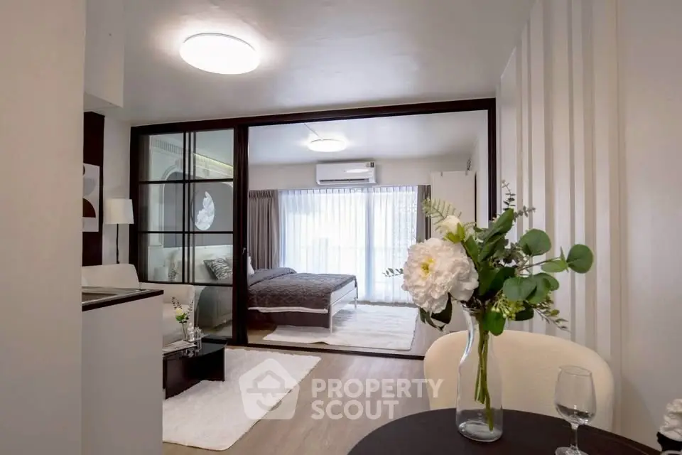 Modern open-plan living room with glass partition leading to a cozy bedroom, featuring elegant decor and natural light.