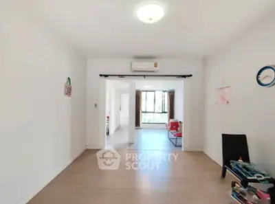 Spacious living room with wooden flooring and air conditioning, leading to a bright area with large windows.