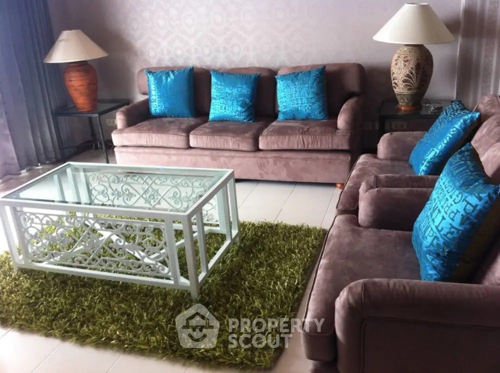 Elegant living room with plush brown sofas, vibrant blue cushions, a unique white coffee table, and a lush green area rug, creating a cozy and stylish space.