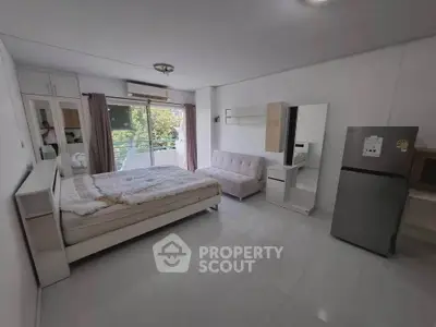 Spacious studio apartment with modern furnishings and balcony view, featuring a cozy bed and sleek refrigerator.