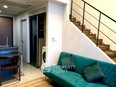 Modern duplex interior with teal sofa, wooden staircase, and compact kitchen featuring washing machine.