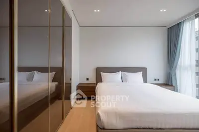 Modern bedroom with sleek design, featuring a large bed, mirrored wardrobe, and natural light from a window.