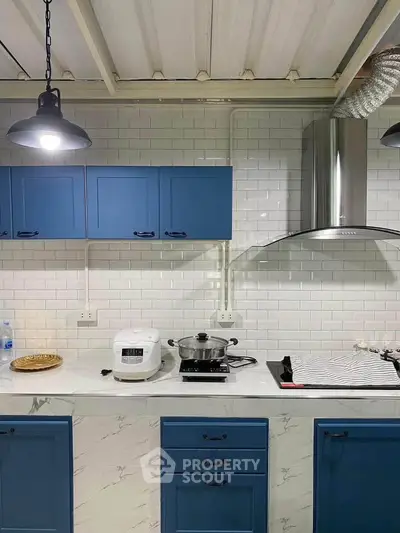 Modern kitchen with blue cabinets, white subway tiles, and marble countertops featuring a rice cooker and stove.