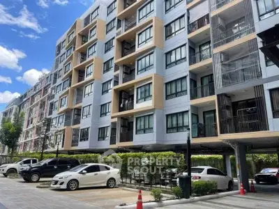 Modern apartment building with parking area and lush greenery under a clear blue sky.