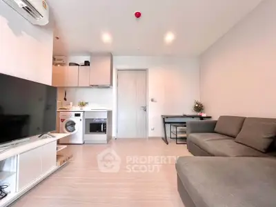 Modern living room with integrated kitchen and washing machine, featuring sleek furniture and bright lighting.