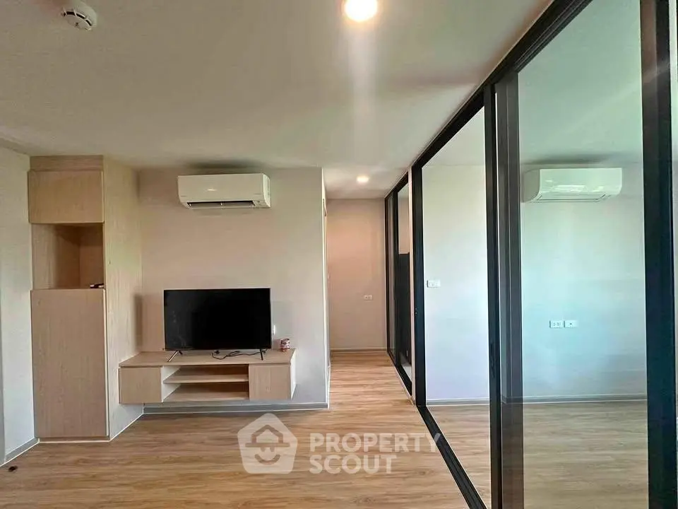 Modern living room with sleek wooden flooring and glass partition, featuring a mounted TV and air conditioning.