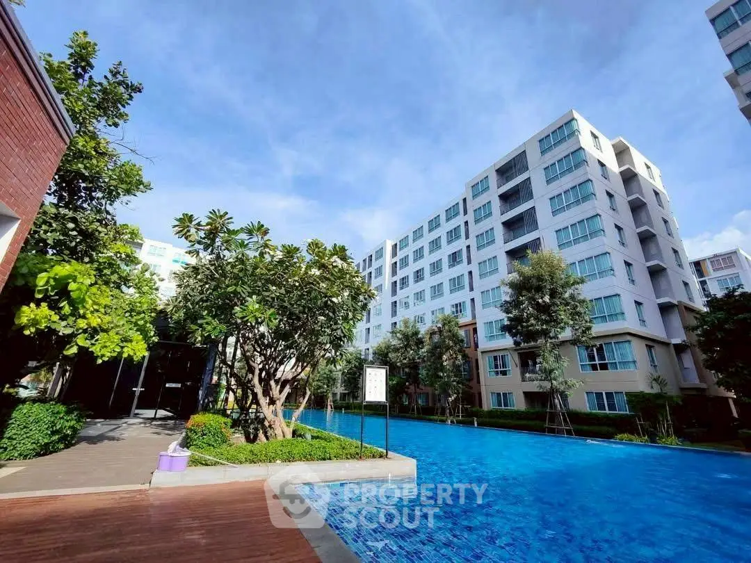 Modern apartment complex with a large swimming pool and lush greenery under a clear blue sky.