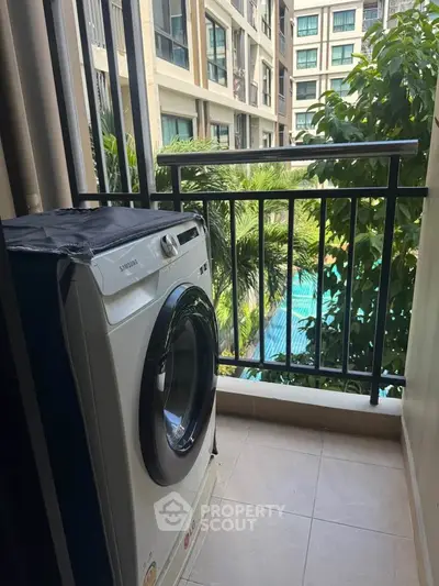 Compact balcony with washing machine overlooking lush greenery and pool view in modern apartment complex.