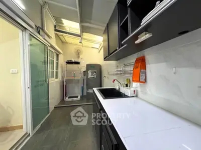 Modern kitchen with sleek black cabinets and white countertops, featuring a washing machine and ample natural light.