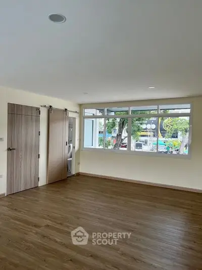 Spacious and well-lit living space with large windows offering an urban view, modern wood flooring, and sliding doors leading to a cozy bedroom.