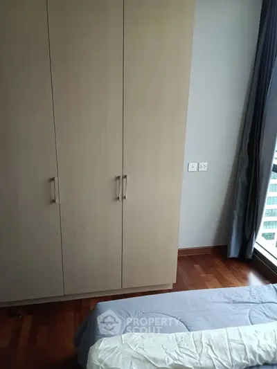 Cozy bedroom with wooden wardrobe and large window offering natural light.