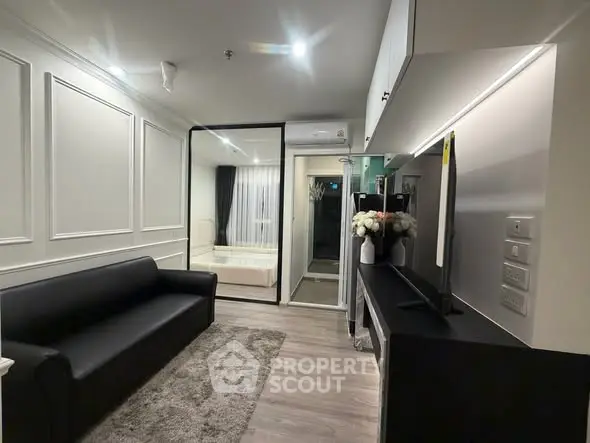Modern living room with sleek black sofa, wall paneling, and large TV, leading to a cozy bedroom.