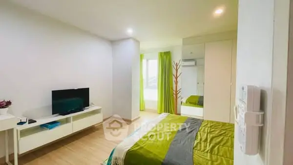 Modern bedroom with green accents and sleek furniture, featuring a wall-mounted TV and ample natural light.