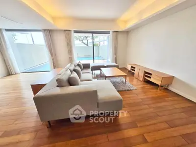 Spacious living room with modern furniture and pool view, featuring wooden flooring and large windows.