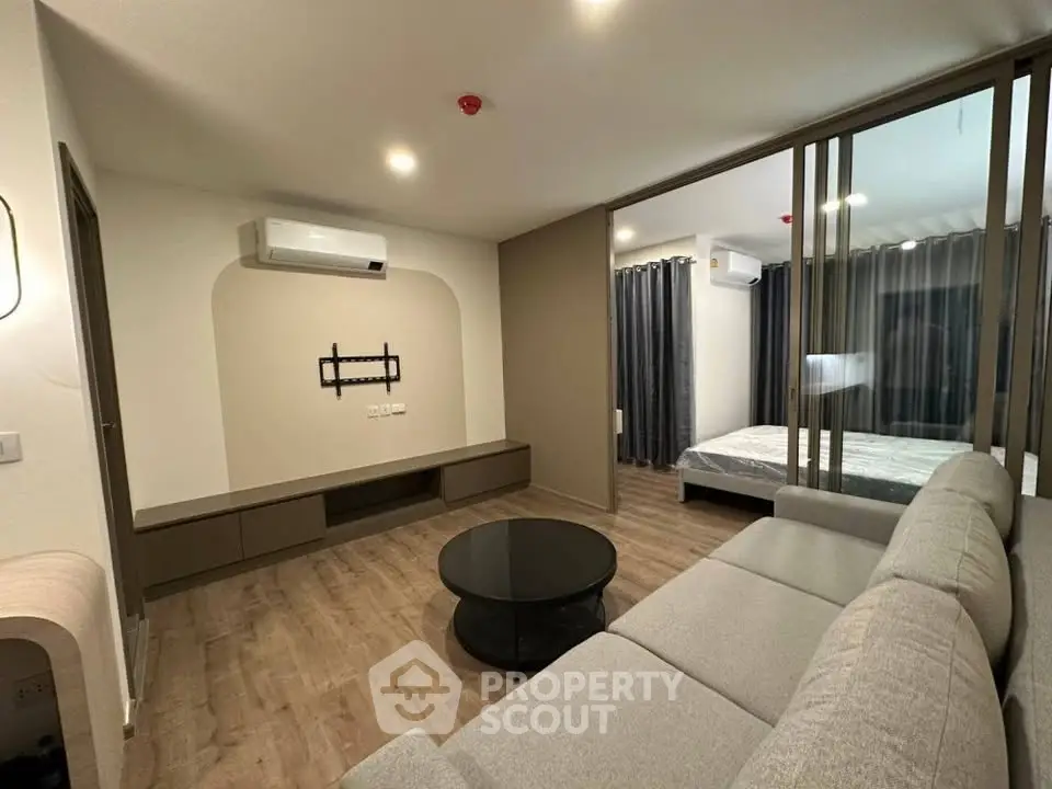 Modern living room with sleek design and glass partition leading to bedroom, featuring air conditioning and stylish furniture.