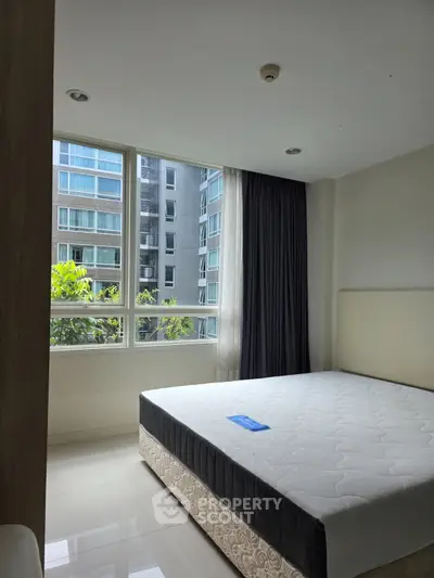 Spacious bedroom with large windows offering a view of modern buildings, featuring a comfortable bed and natural light.