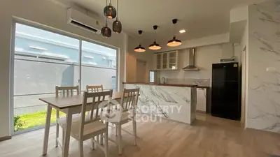 Modern kitchen with marble island and dining area, featuring sleek lighting and large window.