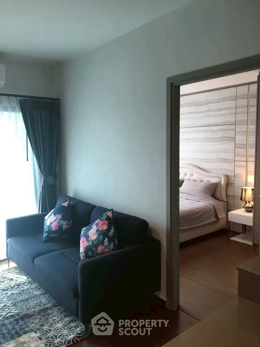 Cozy living room with dark sofa and floral pillows leading to a stylish bedroom with elegant decor.