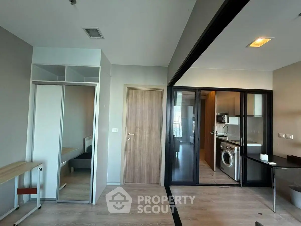 Modern apartment interior with open kitchen and washing machine, featuring sleek wooden flooring and glass partitions.