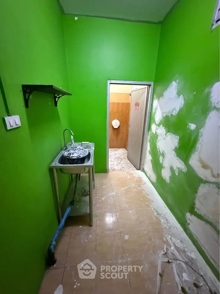 Compact green kitchen with stainless steel sink and open shelving, leading to adjacent room.
