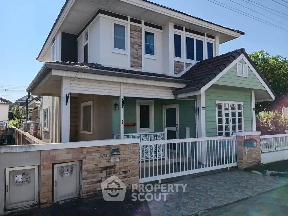 Charming two-story house with green facade and stone accents, featuring a cozy front porch and fenced yard.