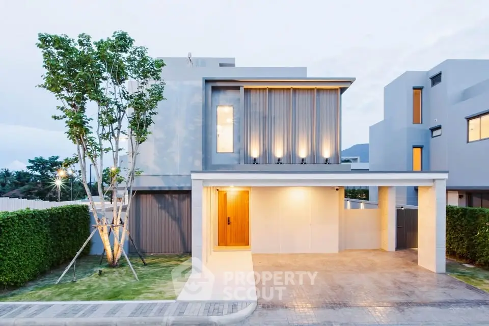 Modern two-story house with sleek design and illuminated facade, featuring a spacious driveway and lush greenery.