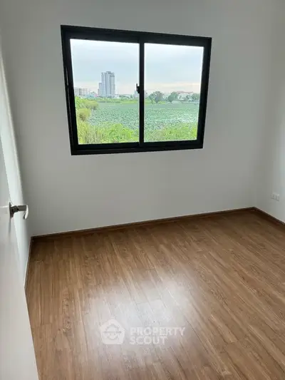 Bright empty room with wooden flooring and large window offering scenic view.