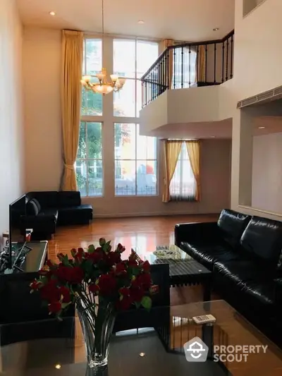 Elegant two-story living space with high ceilings, large windows, and a grand staircase, complemented by sleek black leather sofas and a vase of red roses.