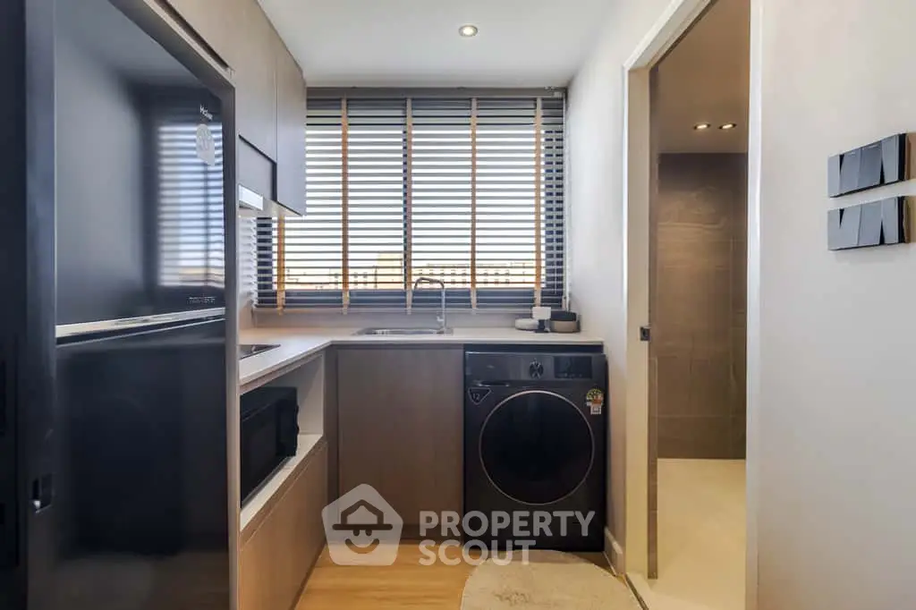 Modern kitchen with sleek appliances and washing machine, featuring wooden cabinetry and large window blinds.