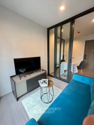 Modern living room with sleek furniture and glass partition, featuring a cozy blue sofa and flat-screen TV.