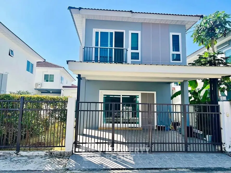 Modern two-story house with balcony and gated driveway in a sunny neighborhood.