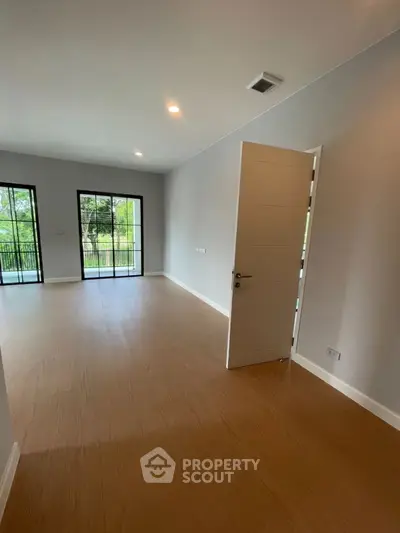 Spacious empty room with wooden flooring and large windows overlooking greenery.