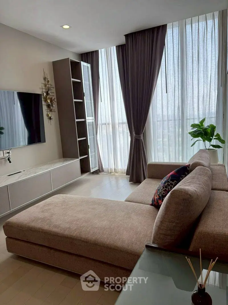 Modern living room with large sofa, wall-mounted TV, and floor-to-ceiling windows with curtains.
