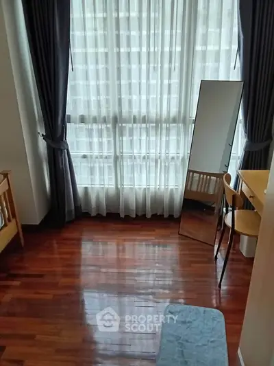 Bright bedroom with large window and wooden flooring, featuring a desk and mirror for a cozy living space.