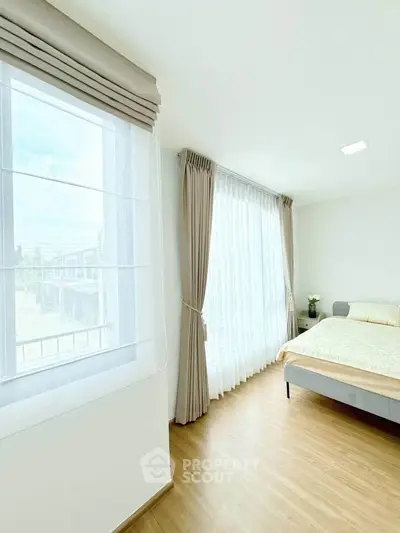 Bright and airy bedroom with large windows and elegant curtains, featuring a cozy bed and wooden flooring.